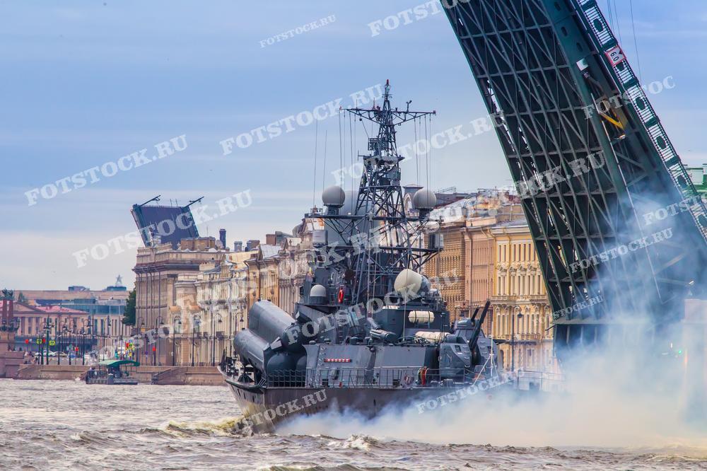 На фотографии запечатлён момент, когда военный корабль проходит под разведённым мостом на реке Неве в Санкт-Петербурге. Фотография подходит для использования в публикациях о военно-морской тематике, истории Санкт-Петербурга или туристических гидах.